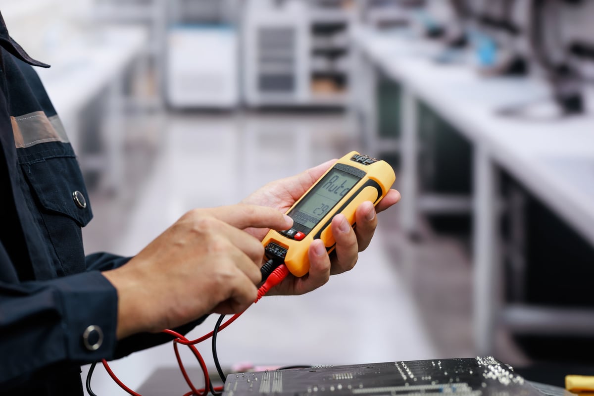 Close-up of a technician using a digital multimeter with test leads to check an electronic circuit board in a workshop.
