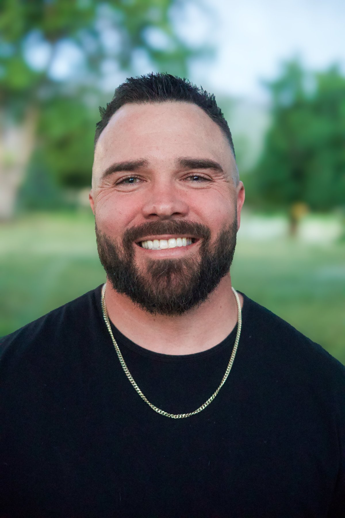 Smiling man with dark hair and beard wearing black t-shirt and chain necklace, outdoors with blurred green trees in background
